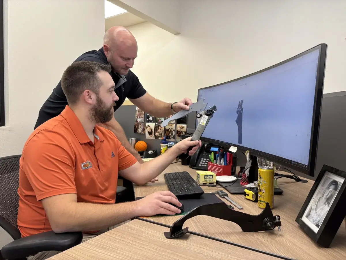 Two mechanical engineers comparing a complex precision mechanical assembly prototype to a 3D CAD model on a computer screen for design validation.